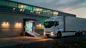 Ein moderner elektrischer Sattelschlepper lädt an einer Logistikhalle mit Solaranlagen, was effiziente Green Logistics symbolisiert.